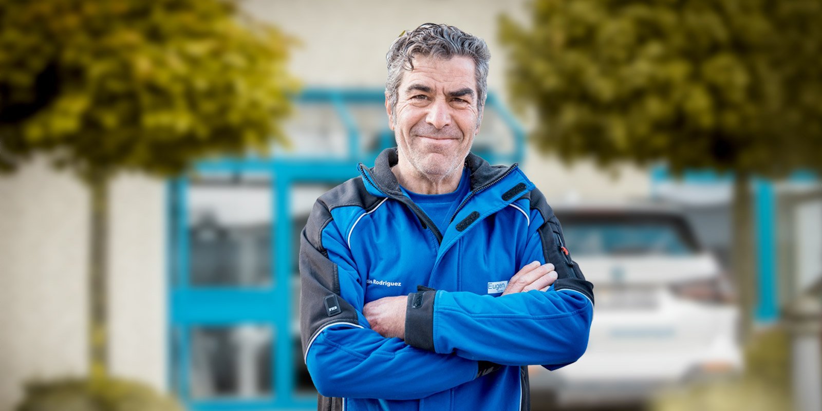 Ein älterer Mann mit grauen Haaren lächelt freundlich, während er mit verschränkten Armen vor einem Gebäude steht.